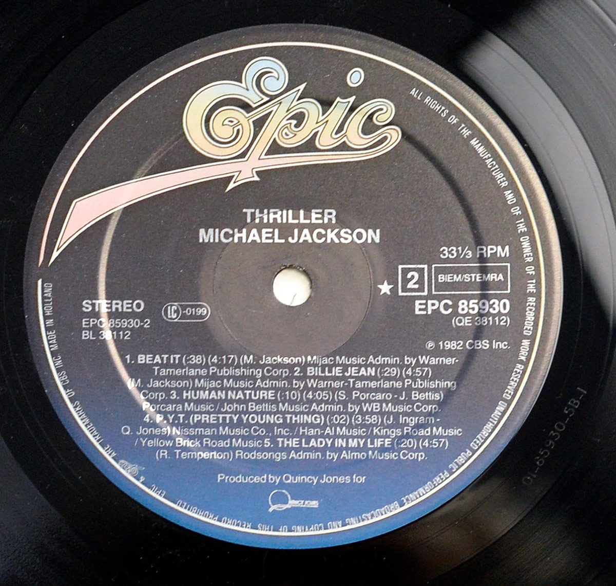 Close-up of the Side Two label from Michael Jackson’s Thriller LP, showing the yellow-and-gold Epic logo at the top above white lettering that reads 'Thriller – Michael Jackson.' The track listing includes the album’s most iconic songs: 'Beat It,' 'Billie Jean,' 'Human Nature,' 'P.Y.T. (Pretty Young Thing),' and 'The Lady in My Life.' The label displays catalog number EPC 85930-2, 33⅓ RPM speed, BIEM/STEMRA rights notice, and the 'Made in Holland' inscription along the rim. The deep blue-to-black gradient background and precise typography epitomize the clean, elegant look of early 1980s European Epic Records pressings.