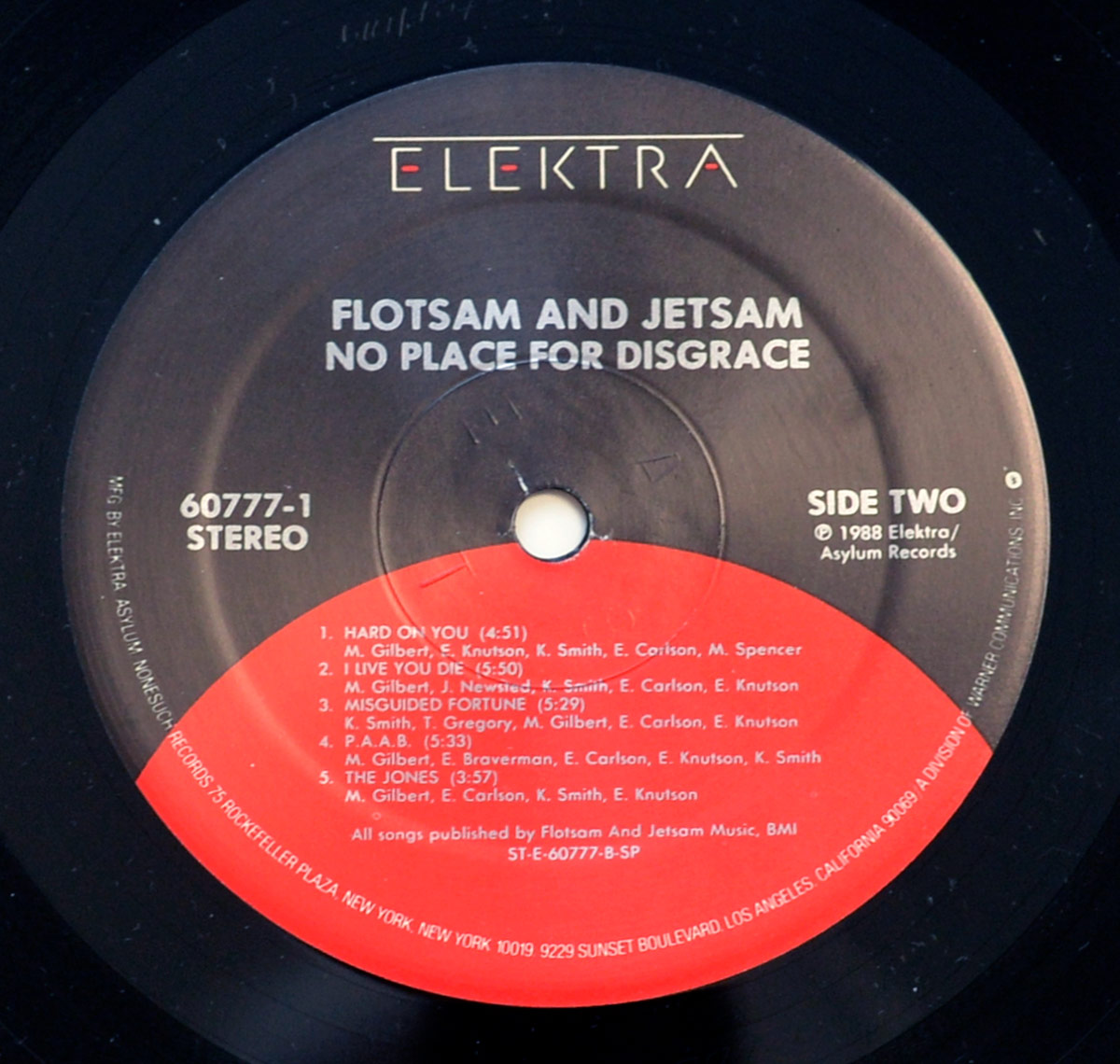 Side Two of Flotsam and Jetsam's 'No Place for Disgrace' vinyl record, released by Elektra/Asylum Records. The label has a black background with the Elektra logo at the top, white text displaying the album and band name, and a red-orange semicircle in the lower half listing tracks and credits for Side Two.