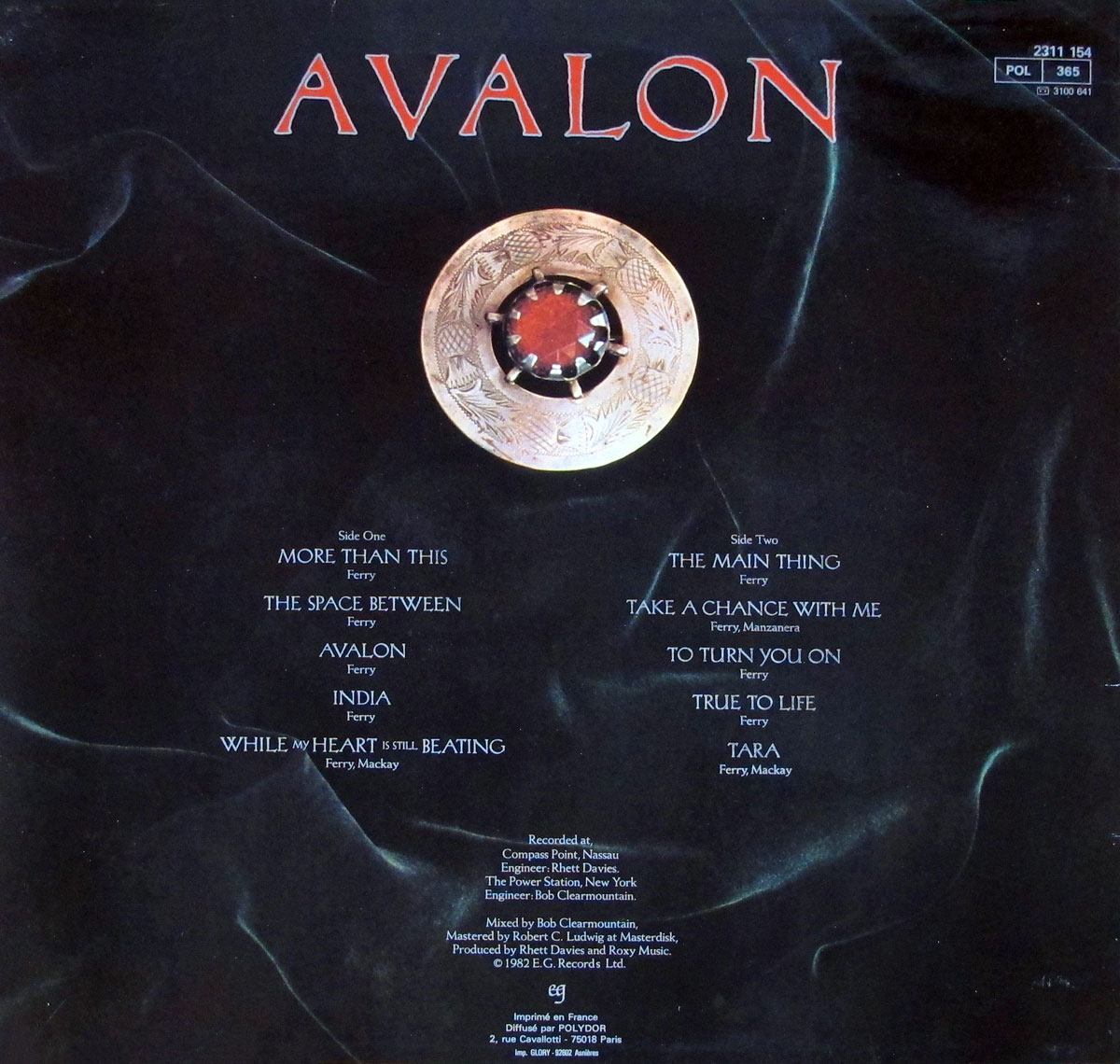 The back cover of Roxy Music's 1982 album Avalon, French release, presents a minimalist layout on a dark velvet-like background. At the top center, the word AVALON appears in bold red uppercase lettering. Beneath it, a circular ornamental medallion with floral engravings and a central deep-red gemstone dominates the design, adding a medieval and regal touch. On either side of the medallion, the track listing for Side One and Side Two is arranged in elegant serif font, with songwriter credits (Ferry, Mackay, Manzanera) carefully aligned beneath each title. At the bottom, production credits list Compass Point Studios, The Power Station, engineers Rhett Davies and Bob Clearmountain, mastering by Robert C. Ludwig, and producers Rhett Davies and Roxy Music. The catalog number 2311 154 appears in small type in the upper right corner, with Polydor distribution notes and French imprint details at the bottom.