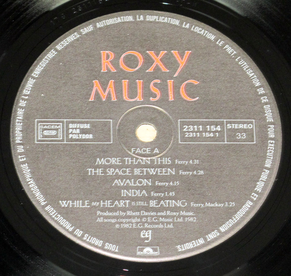 Close-up of the Side One record label for Roxy Music's Avalon, France release (1982). The label is dark gray with red 'ROXY MUSIC' printed in bold capitals across the top. The center hole is surrounded by circular etching lines. To the left are SACEM and Polydor distribution marks, while to the right the catalog number 2311 154, matrix code 2311 154 1, and STEREO 33 format details are shown. Beneath the center, the tracklist for Side One is displayed in serif white type: 'More Than This' (4:31), 'The Space Between' (4:28), 'Avalon' (4:15), 'India' (1:45), and 'While My Heart Is Still Beating' (3:25). Credits at the bottom note producers Rhett Davies and Roxy Music, copyright to E.G. Music Ltd 1982, and Polydor imprint. The fine circular text along the edge states legal restrictions in French. The overall design is formal, symmetrical, and functional, ensuring legibility.