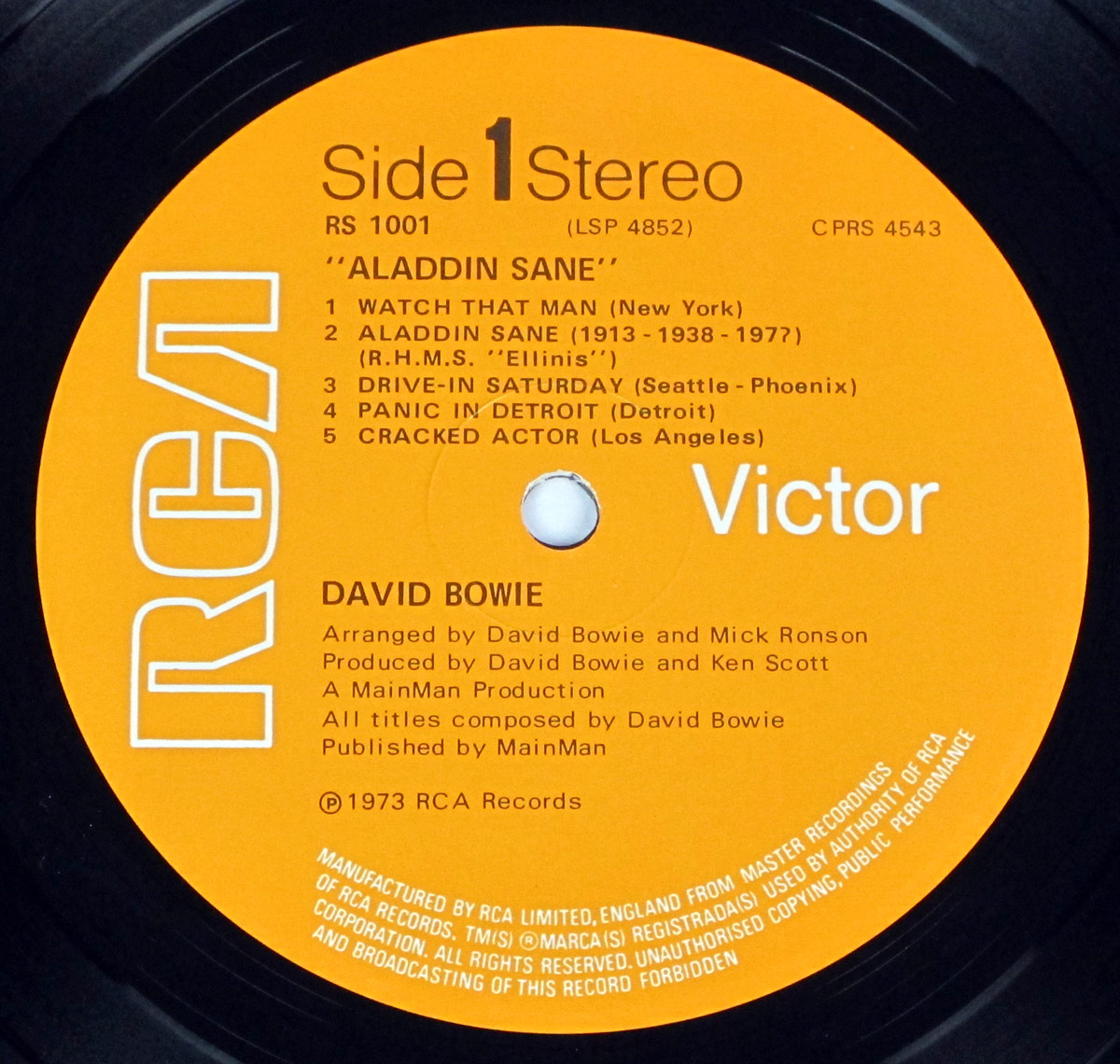 Side 1 record label of Aladdin Sane by David Bowie, released on RCA Victor in 1973. The label features a bold orange background with the large RCA logo displayed vertically on the left and the Victor logo in white on the right. The album title “ALADDIN SANE” is printed in capital letters, followed by the tracklist for Side 1, which includes five songs along with their respective locations. Below the tracklist, production credits mention David Bowie, Mick Ronson, and Ken Scott. The bottom of the label includes copyright information and a disclaimer about unauthorized copying and broadcasting. The center hole is precisely cut, with record grooves faintly visible around the edges.