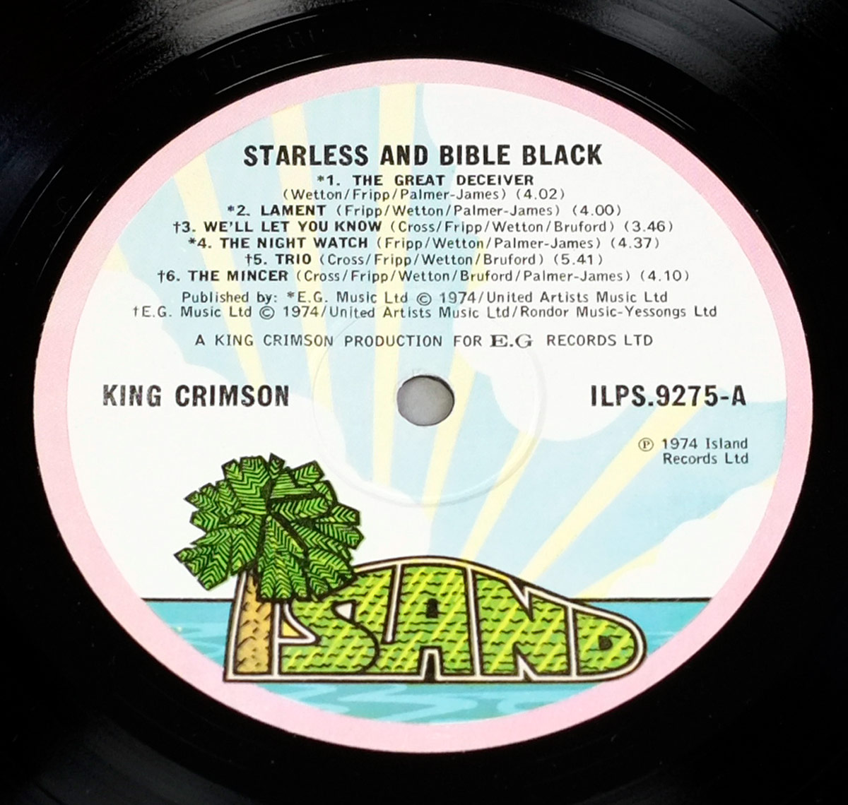 Close-up of the Side One record label for King Crimson’s 'Starless and Bible Black' (1974). The label features Island Records’ iconic 'Pink Rim / Palm Tree' design: a hand-drawn tropical island with a green palm tree, brown trunk, and the bold word ISLAND in textured green letters rising from turquoise-blue sea and yellow sun rays. The text area above lists all six tracks with songwriting credits and publishing details. Printed in England with catalogue number ILPS.9275-A, this pressing is the 1974 UK edition released by Island Records for E.G. Records Ltd.