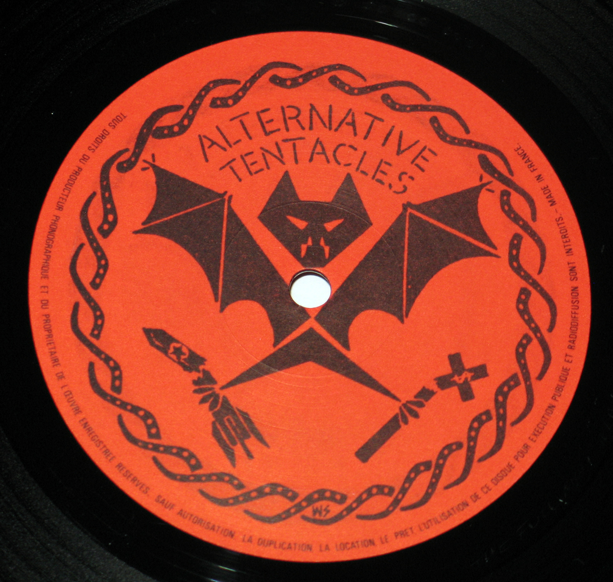 Record label of Dead Kennedys’ Frankenchrist featuring the Alternative Tentacles logo. The design includes a black stylized bat with a fanged skull, crossed symbols of a rocket and a cross, and a coiled serpent-like border on a red-orange background. The text ‘Alternative Tentacles’ arches over the bat, reinforcing the underground punk label’s rebellious and subversive aesthetic.