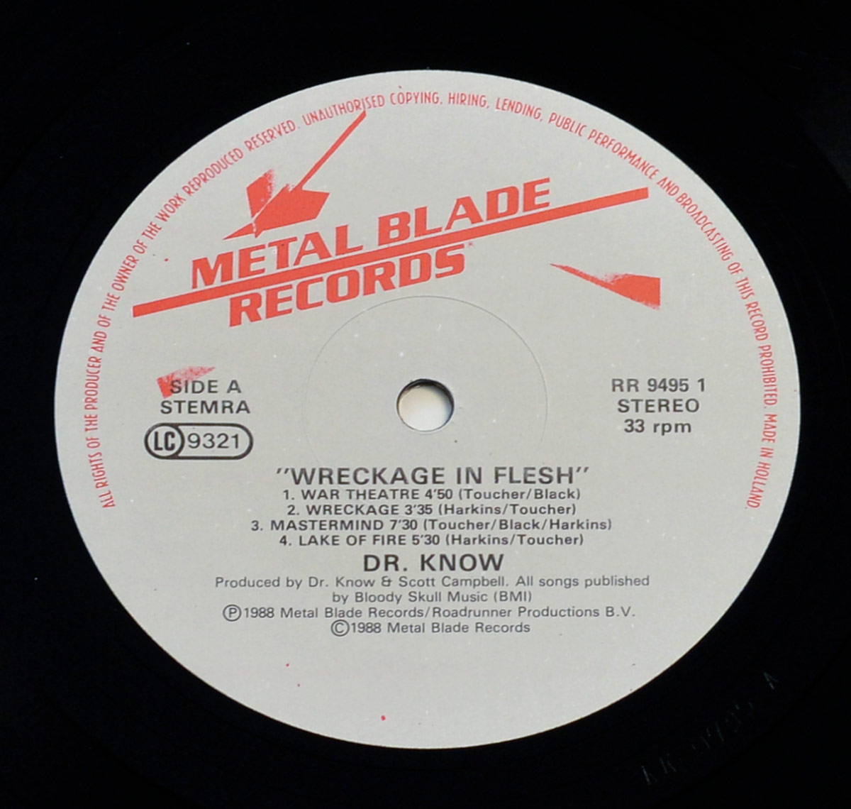 Close-up of Side One record label of 'Wreckage in Flesh' by Dr. Know. The label is off-white with red 'Metal Blade Records' branding across the top, including the logo with a stylized blade. It lists four tracks: 'War Theatre,' 'Wreckage,' 'Mastermind,' and 'Lake of Fire,' along with their durations and songwriting credits. The bottom section credits production to Dr. Know and Scott Campbell, published under Bloody Skull Music (BMI). The catalog number 'RR 9495 1' and 'Stereo 33 rpm' are also printed. A circular hole is centered for mounting on a turntable. The perimeter of the label states copyright information and restrictions. Visit https://vinyl-records.nl