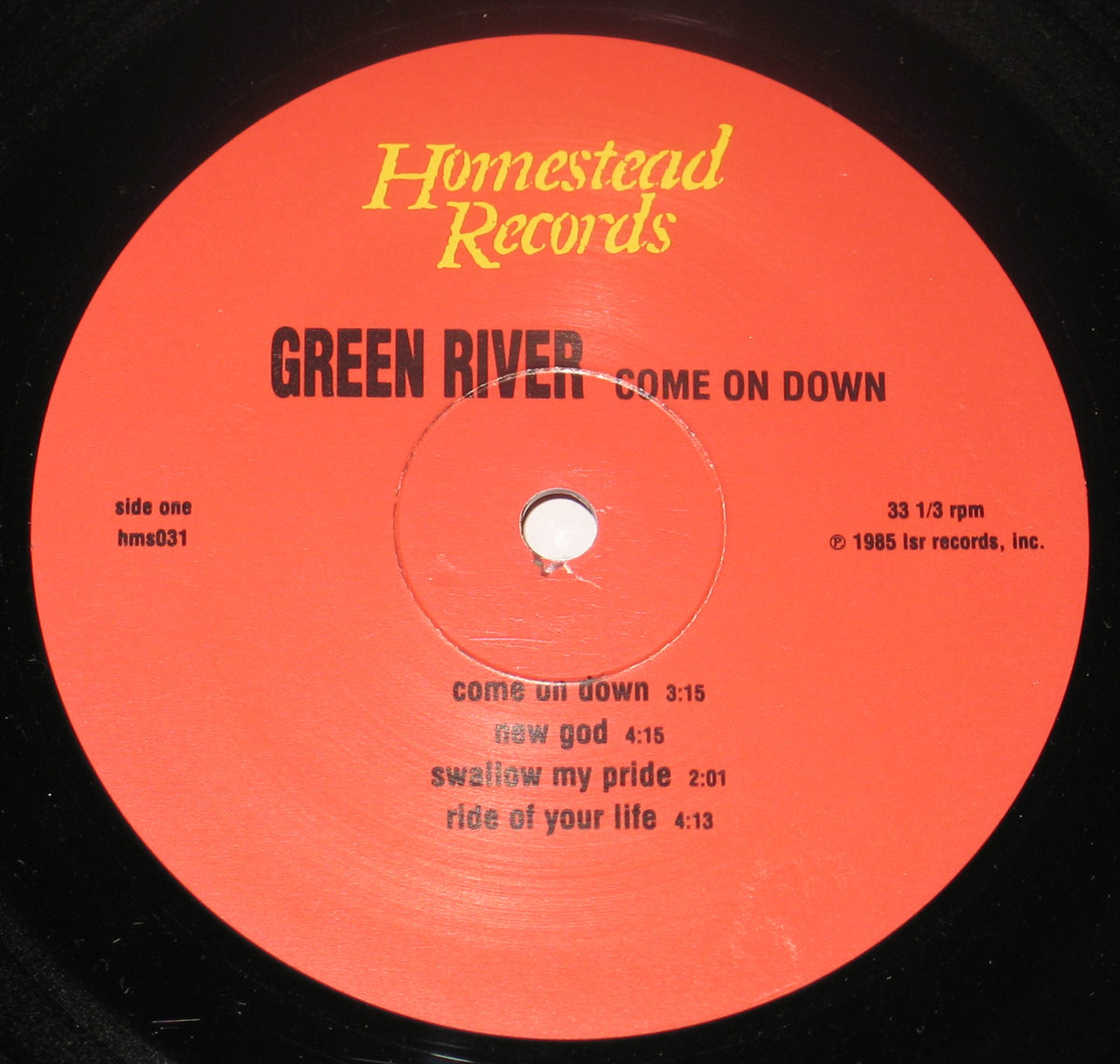 Vinyl record label of Green River's 'Come On Down'. The label is a bold red-orange color with black and yellow text. 'Homestead Records' is displayed in stylized yellow font at the top, with 'GREEN RIVER – COME ON DOWN' in bold black capital letters below. The tracklist for Side One is listed, including 'Come On Down,' 'New God,' 'Swallow My Pride,' and 'Ride of Your Life.' The label also indicates the catalog number 'hms031,' playback speed of 33 1/3 RPM, and copyright details from 1985.