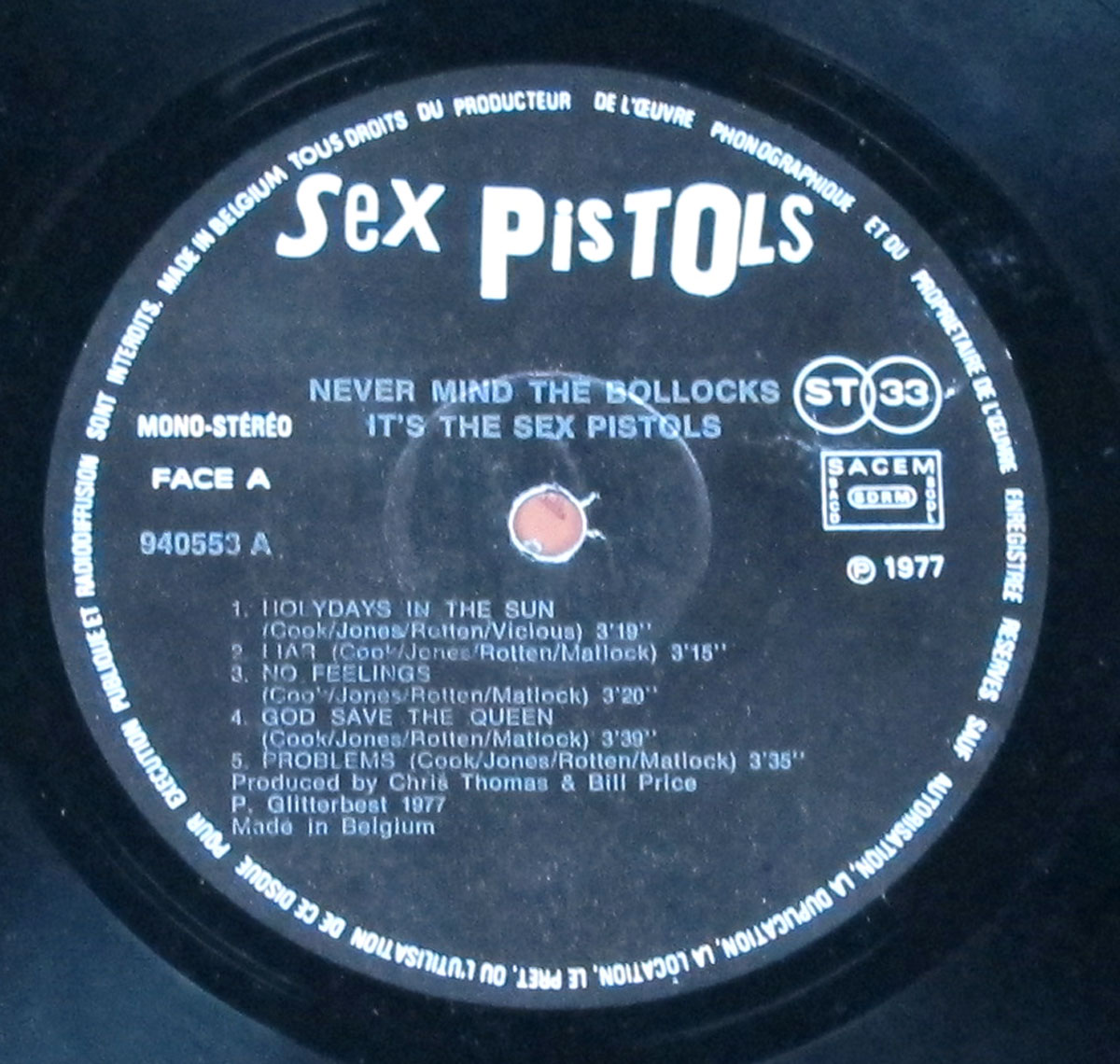 Close-up of the Side A record label from the Belgium export for France edition of Never Mind the Bollocks. Black paper label with white “Sex PisTOLS” at the top, French text ring around the edge, and centered album title “NEVER MIND THE BOLLOCKS IT’S THE SEX PISTOLS.” Left side reads MONO-STÉRÉO and FACE A; catalog number 940553 A appears mid-left. To the right are twin ST 33 logos and a boxed SACEM rights logo. Track list shows a notable typo: HOLYDAYS IN THE SUN, followed by I AM, NO FEELINGS, GOD SAVE THE QUEEN, PROBLEMS with timings. Footer credits: Produced by Chris Thomas & Bill Price, ℗ Glitterbest 1977, Made in Belgium.