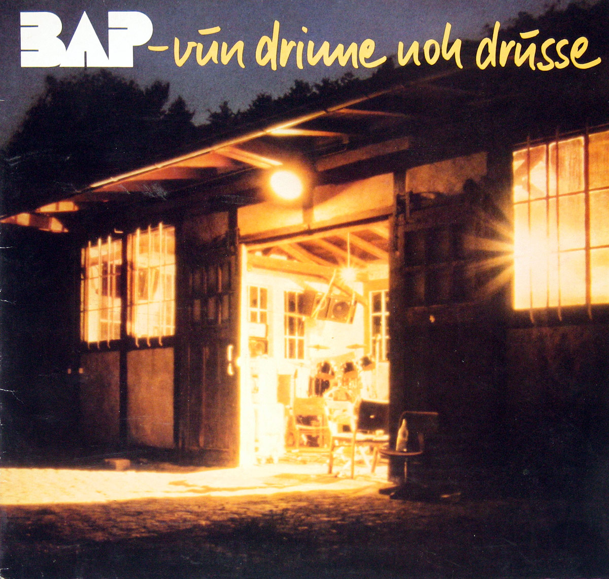 The front cover of BAP's 1982 album 'Vun Drinne Noh Drusse' shows a warmly lit rehearsal space inside a rustic wooden barn, viewed from outside at dusk.