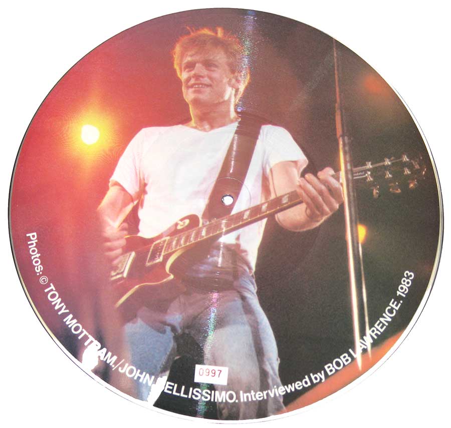 Bryan Adams smiling in white t-shirt, holding a black electric guitar on stage, bathed in warm stage lights. Picture disc from 1983, photo by Mottram and Bellissimo.