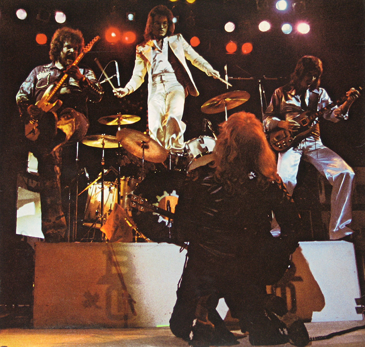 Live stage photo of BTO with members playing guitar, bass, and drums under dramatic lighting; one member kneeling, another elevated above the drum kit