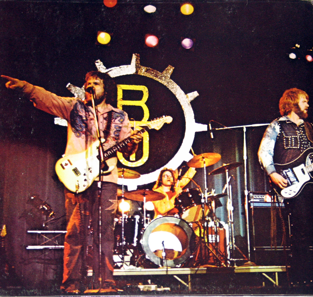 Live stage photo of Bachman Turner Overdrive performing in front of a giant metallic gear logo with BTO initials, spotlighted and energized