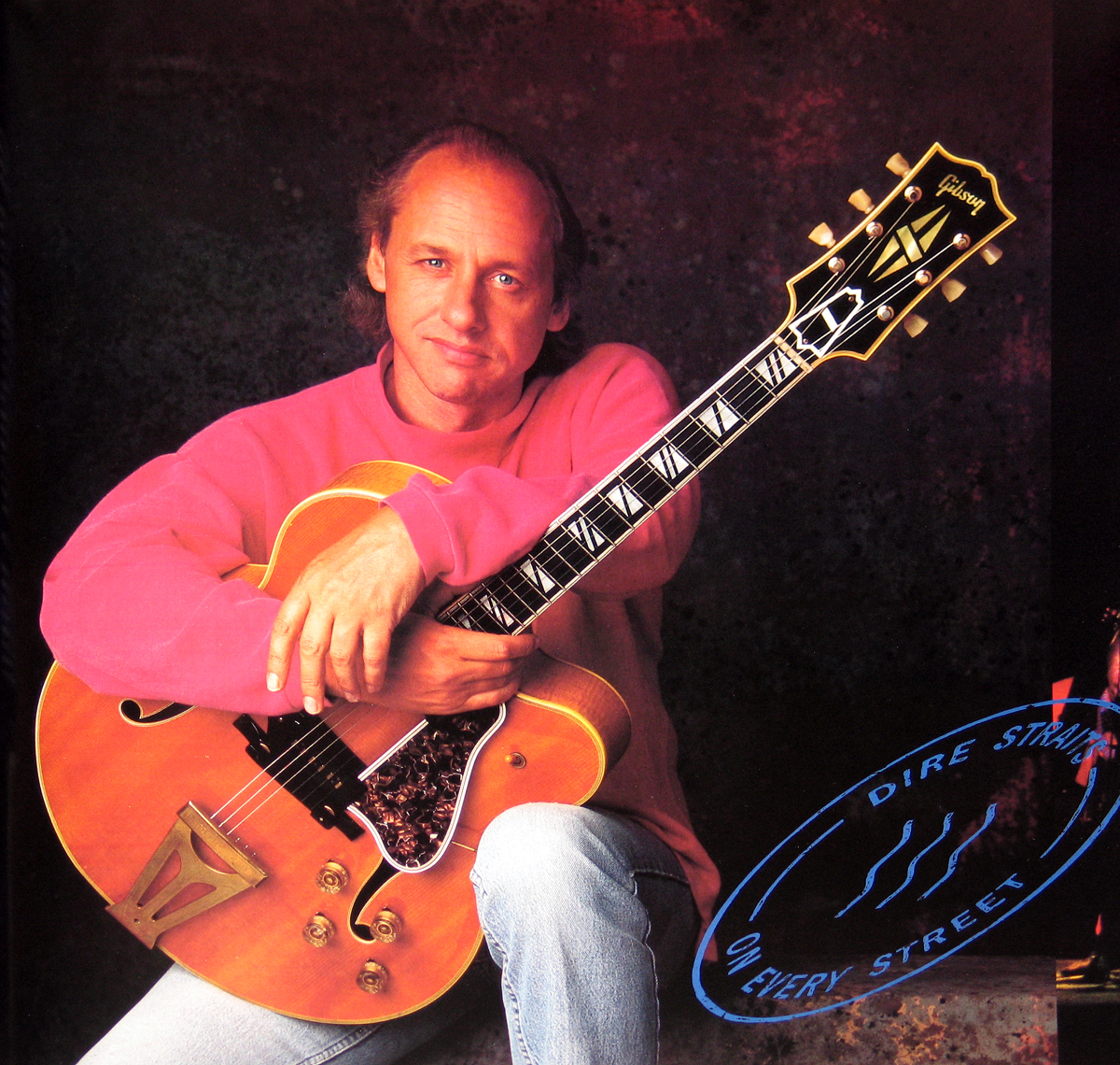 Full-page inner sleeve photograph from Dire Straits’ 1991 album On Every Street showing Mark Knopfler seated casually with a natural wood-finish Gibson archtop guitar. He wears a soft pink sweatshirt and light denim jeans, his arm resting across the guitar’s body with quiet confidence. The lighting is warm, creating a contrast between his vivid clothing and the dark marbled studio backdrop. The iconic blue ‘Dire Straits On Every Street’ postal stamp emblem appears in the lower right corner, uniting the design theme of the album. The image reflects the intimacy and craftsmanship that defined Knopfler’s tone and composure during the band’s final studio era.