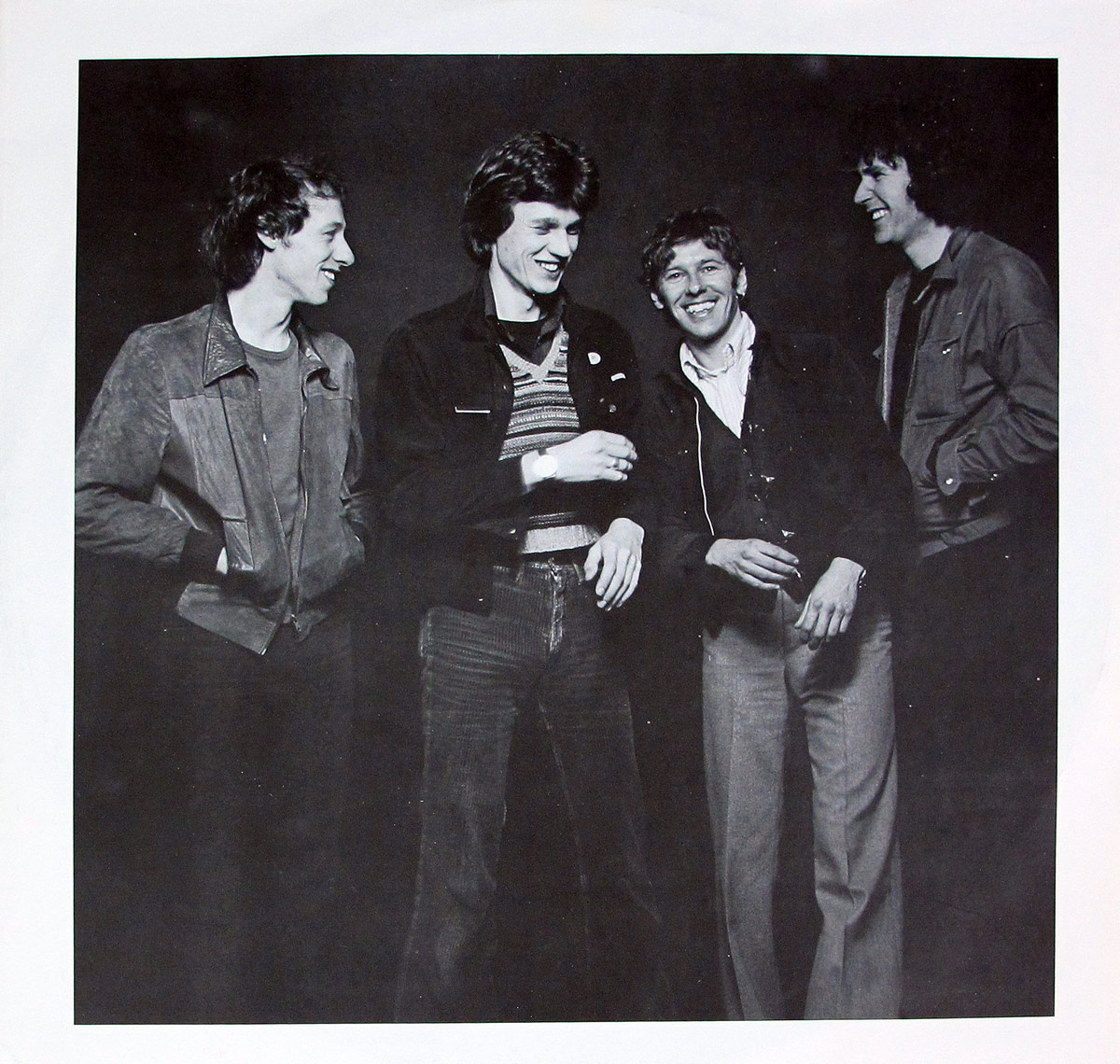 Black-and-white inner sleeve photograph of Dire Straits from the French Black Vertigo 2nd Pressing of their self-titled debut album. All four band members—Mark Knopfler, David Knopfler, John Illsley, and Pick Withers—stand together in a relaxed, informal pose against a dark studio backdrop. Mark Knopfler is on the far left with hands in pockets, smiling toward David Knopfler, who stands beside him laughing with his jacket open and striped sweater visible. John Illsley leans forward slightly, grinning, while Pick Withers on the far right laughs openly. Their casual clothing, natural expressions, and unpolished stance evoke the raw charm of a young band on the rise, captured in a candid, almost spontaneous moment that contrasts the minimalist design of the album’s exterior.