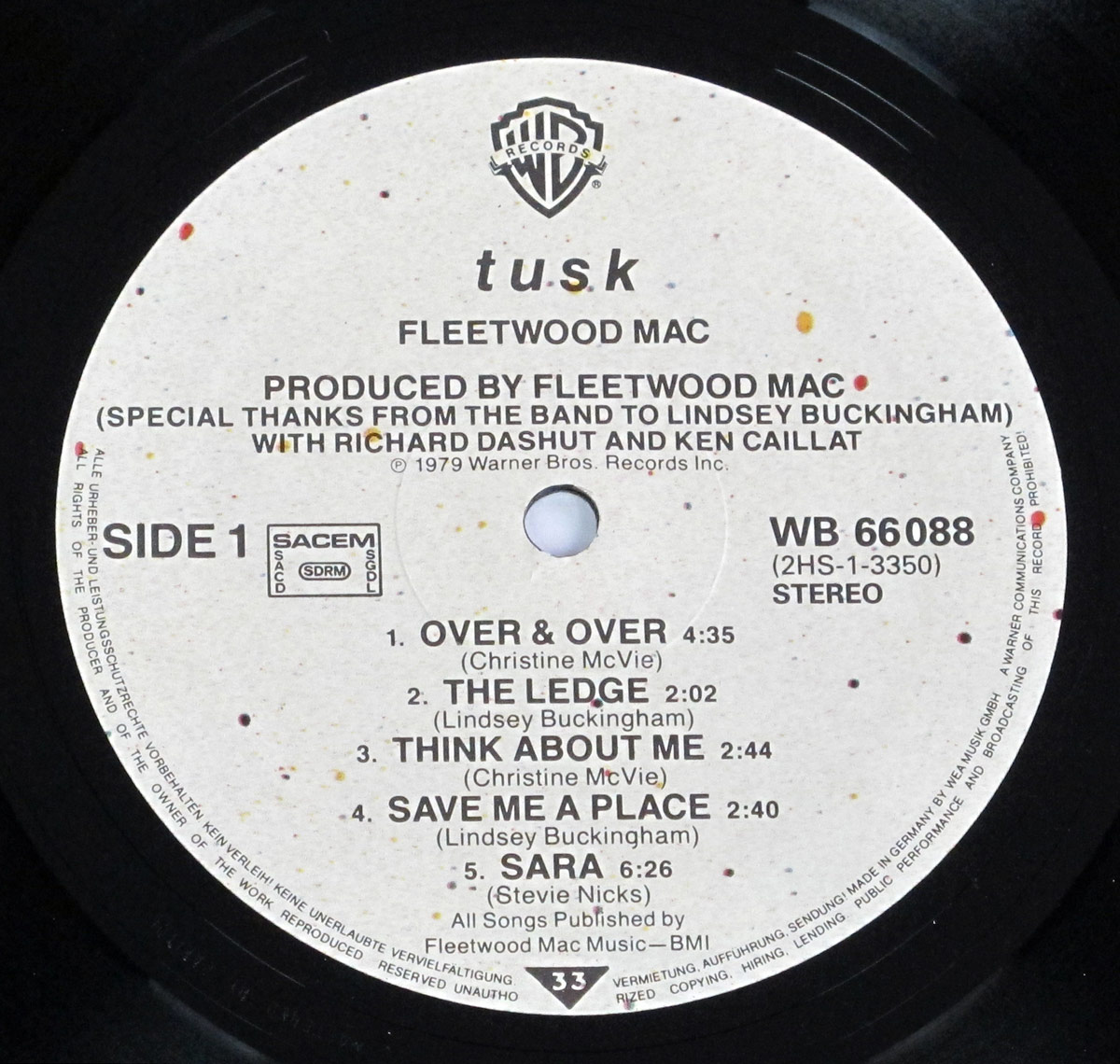 Close-up of Fleetwood Mac's 'Tusk' vinyl record label, pressed on a speckled off-white background with red, yellow, and black flecks. The Warner Bros. Records logo is centered at the top, followed by the album title, band name, and production credits. The tracklist for Side 1 is printed in bold black text, detailing five songs with their durations and songwriter credits.