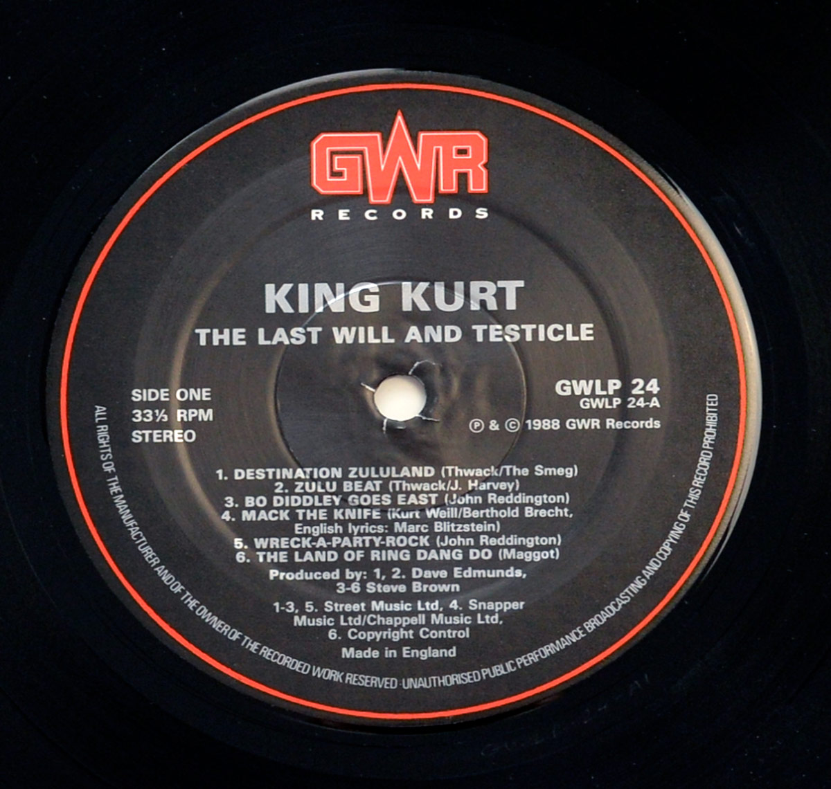 Close-up of Side One record label from King Kurt's 1988 UK psychobilly album The Last Will and Testicle, released on GWR Records GWLP 24. The black label features the bold red GWR Records logo at the top. Centered in white text are the band name King Kurt and the album title. The left side lists Side One, 33⅓ RPM, Stereo. The track listing includes Destination Zululand (Thwack/The Smeg), Zulu Beat (Thwack/J. Harvey), Bo Diddley Goes East (John Reddington), Mack the Knife (Kurt Weill/Berthold Brecht, English lyrics Marc Blitzstein), Wreck-a-Party-Rock (John Reddington), and The Land of Ring Dang Do (Maggot). Producers include Dave Edmunds and Steve Brown. Publishing details credit Street Music Ltd, Snapper Music Ltd, Chappell Music Ltd, and Copyright Control. Around the outer rim is a copyright and rights statement, with 'Made in England' printed at the bottom.