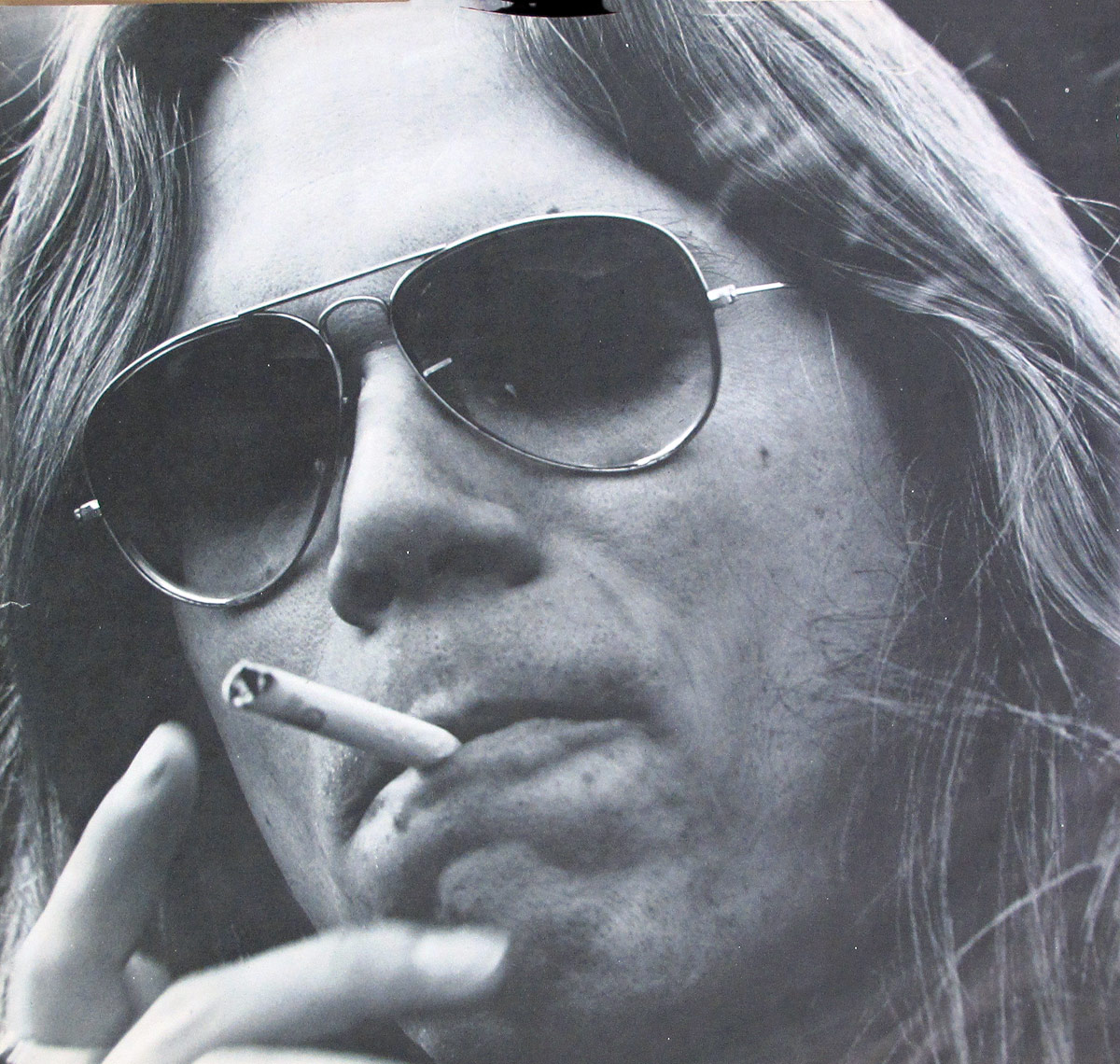 Black and white close-up portrait of Peter Kaukonen from the original custom inner sleeve of 'Black Kangaroo' album. The musician is wearing aviator sunglasses and has long, flowing hair. He holds a cigarette between his lips while his fingers rest near his mouth. The grainy texture and dramatic contrast in the photograph add a raw and rebellious energy, reflecting the rock ‘n’ roll aesthetic of the album. The intense gaze behind the reflective sunglasses enhances the mystique and attitude of the image.