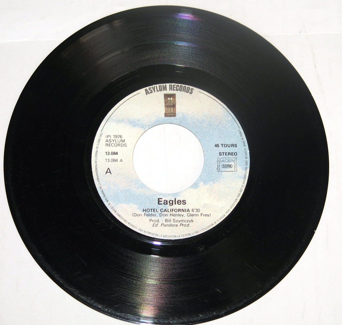 7-inch vinyl single of “Hotel California” by the Eagles, released by Asylum Records. The label has a light blue and white sky-like gradient background with the Asylum Records logo at the top. The text includes the band’s name, “Eagles,” in bold black letters, followed by the song title, “Hotel California,” and its duration of 6 minutes and 30 seconds. Songwriters Don Felder, Don Henley, and Glenn Frey are credited, along with producer Bill Szymczyk. The label also displays the catalog number “13.084 / 13.084 A,” the “45 Tours” (45 RPM) format, and rights society markings such as SACEM and GEMA, indicating a European pressing.