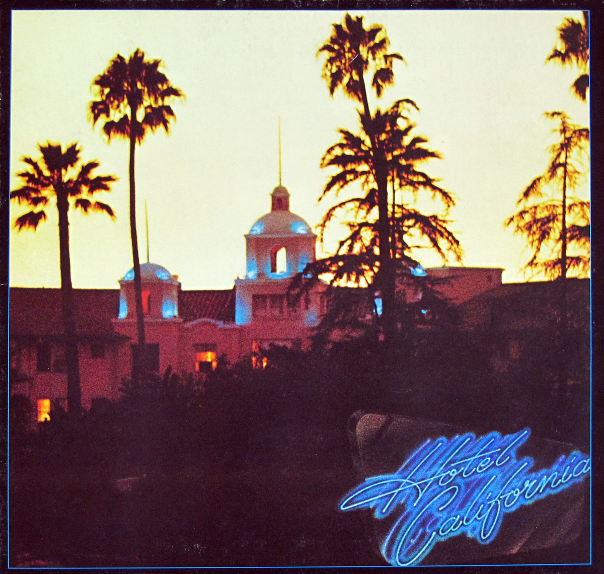 ‘Album cover of The Eagles’ Hotel California, featuring a dusk photograph of the Beverly Hills Hotel with silhouetted palm trees against a warm yellow-orange sky. The hotel building is softly illuminated, with faintly glowing arches and domes. In the lower right corner, the album title “Hotel California” is displayed in blue cursive neon, adding a surreal, dreamlike quality to the image.’