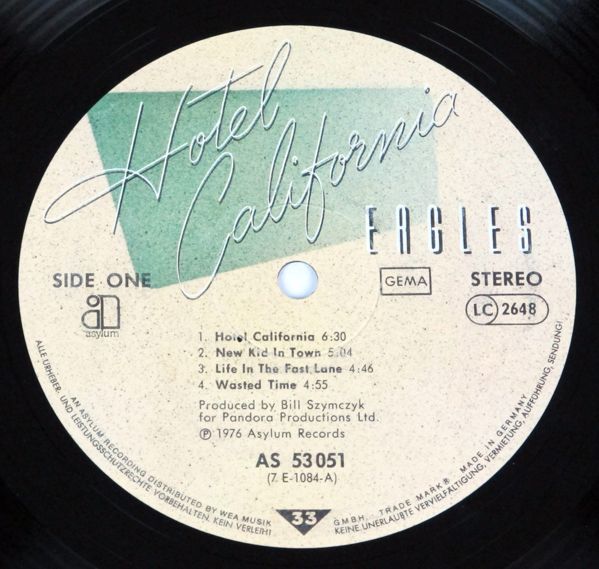 A close-up of the vinyl record label for the Eagles’ 1976 album Hotel California, German release. The label has a beige background with a green banner behind the album title, written in elegant cursive. ‘Eagles’ is displayed in bold capital letters. It includes track listings for Side One, production credits, and distribution details from Asylum Records, marked with ‘Made in Germany’ and GEMA licensing.