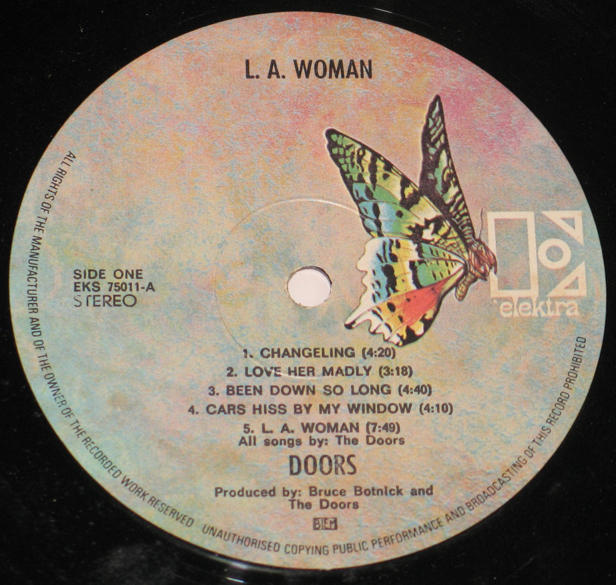 Close-up of the Elektra record label for The Doors' album L.A. Woman, Side One. The circular label has a textured pastel background blending blue, pink, and gray tones. On the right side is the Elektra butterfly logo in green, yellow, and red, next to the geometric Elektra wordmark in white. The top center reads L.A. Woman. On the left side, it indicates Side One with catalog number EKS 75011-A and Stereo. The track listing is printed in black with five songs: Changeling, Love Her Madly, Been Down So Long, Cars Hiss By My Window, and L.A. Woman. The bottom credits show all songs by The Doors, produced by Bruce Botnick and The Doors, with the rights society logo BIEM.