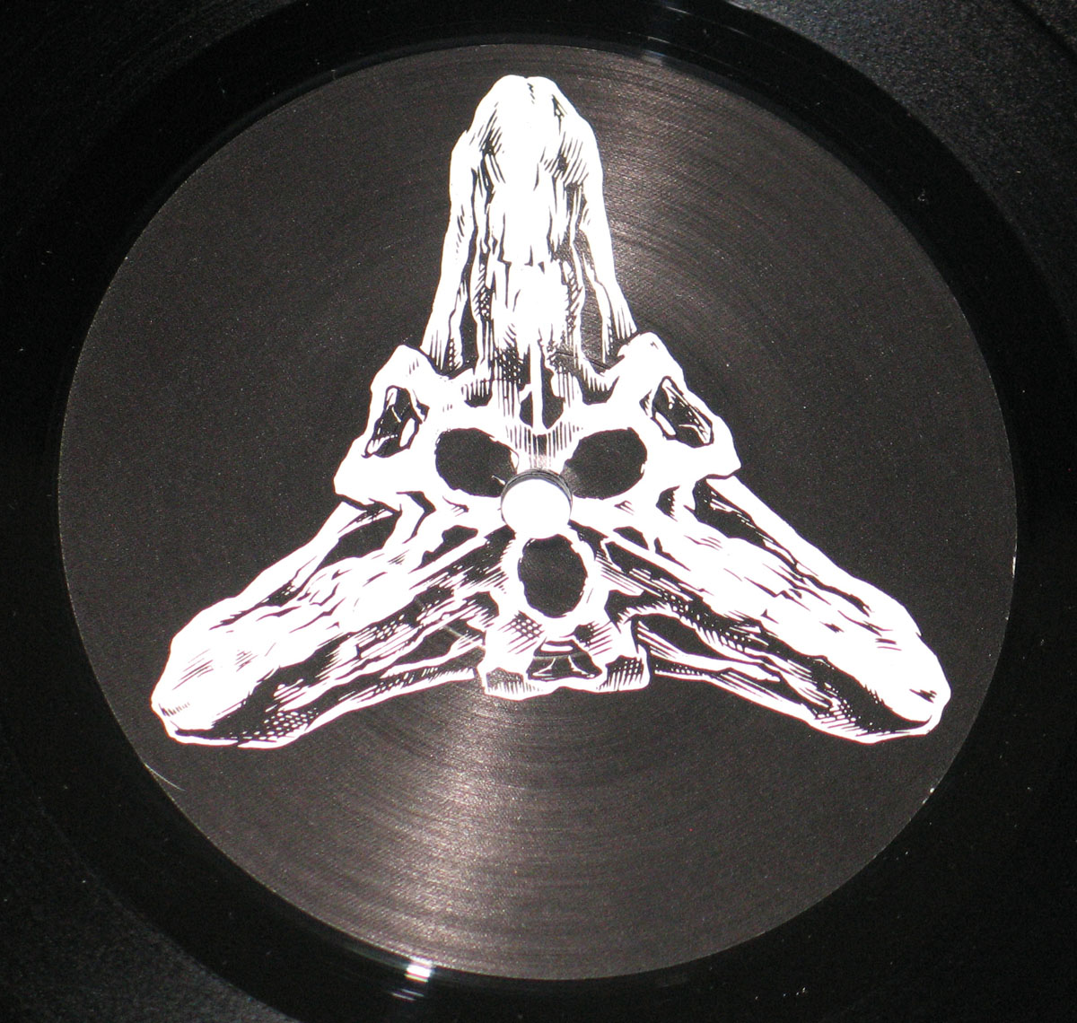 The record label artwork displays a stark, skeletal design rendered in high-contrast black and white. It features an abstract, three-pronged bone structure that forms a symmetrical, almost triangular shape. At the top, a haunting, elongated figure is draped over the central peak, with its form blending seamlessly into the skeletal elements below. The intricate detailing and negative space within the structure give it a dark, visceral quality, with hollowed areas adding depth to the skeletal motif. The design is simple yet striking, evoking themes of death and decay in line with the album’s overall aesthetic.