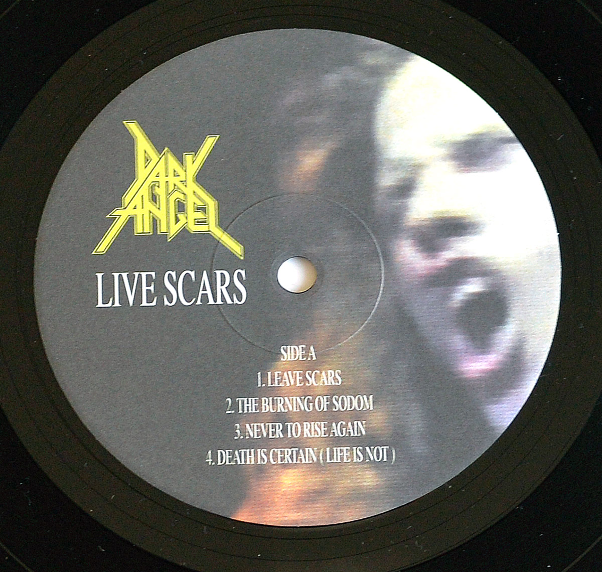Side A label of Dark Angel's 'Live Scars' 12-inch vinyl record. The label features a high-contrast, blurred close-up of vocalist Ron Rinehart screaming. The band's yellow logo is positioned in the upper left, with 'Live Scars' in bold white text below it. The tracklist for Side A is centered in white serif font.