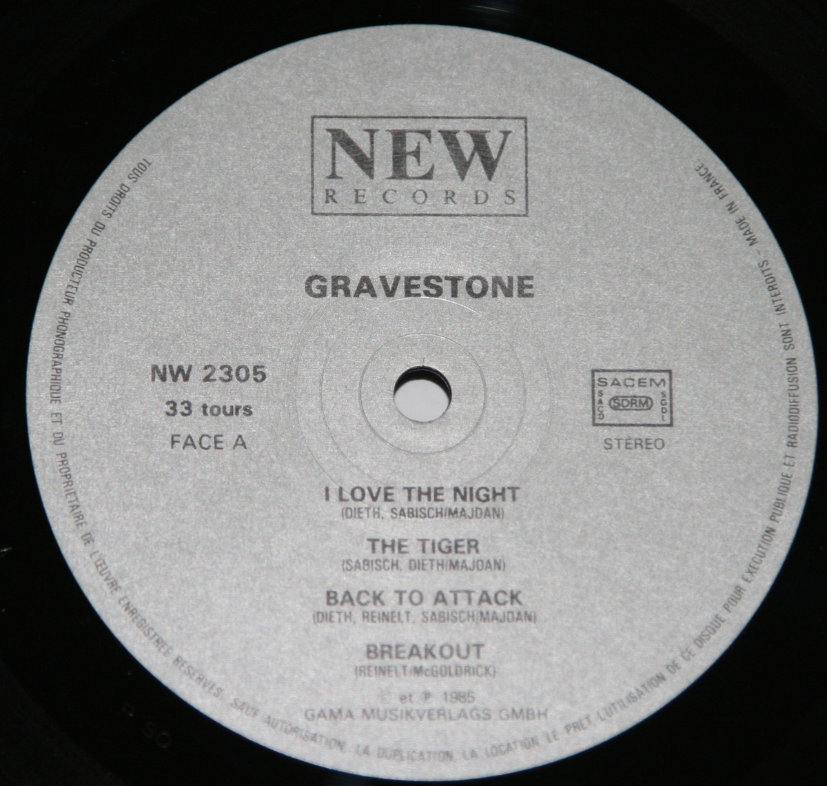 Close-up of Side A record label for Gravestone’s 1985 album Back to Attack. The label design is minimalist and clean, printed on a matte silver background with black lettering. At the top appears the NEW RECORDS logo inside a simple rectangular frame. Beneath it, the band name GRAVESTONE is centered in bold capitals, followed by the catalog number NW 2305, playback speed 33 tours, and designation FACE A. The four track titles are neatly aligned: I Love the Night, The Tiger, Back to Attack, and Breakout, each credited to the band’s composers Dieth, Sabisch, Reinelt, Majdan, and McGoldrick. To the right, the SACEM rights box is visible, and below, the text ‘GAMA Musikverlags GmbH © 1985’ confirms the pressing’s German origin. Around the rim, small French text denotes copyright protection and reproduction restrictions, typical of European metal LP issues from the mid-1980s.