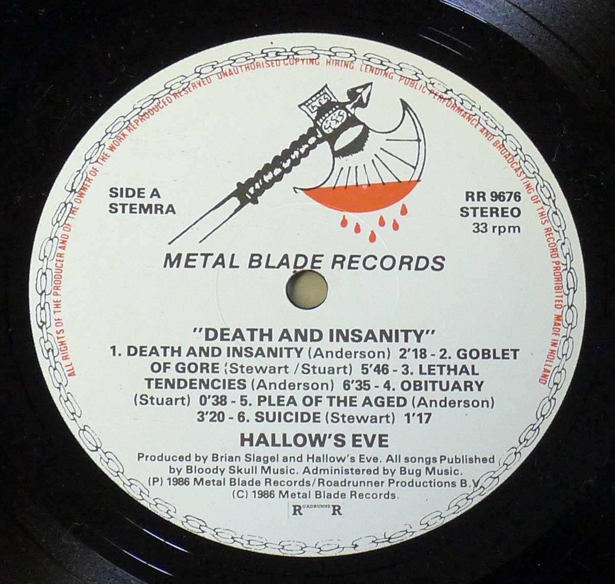 Close-up of the Side A record label for Hallows Eve's Death and Insanity. The label features the Metal Blade Records logo, consisting of a battle axe dripping with blood. The text is printed in black on a white background with a chain-like border around the edge. The album title, track listing, and credits, including the producer Brian Slagel and the publishing information for Bloody Skull Music, are clearly visible. The catalog number RR 9676 is printed along with the stereo and 33 rpm format specifications.