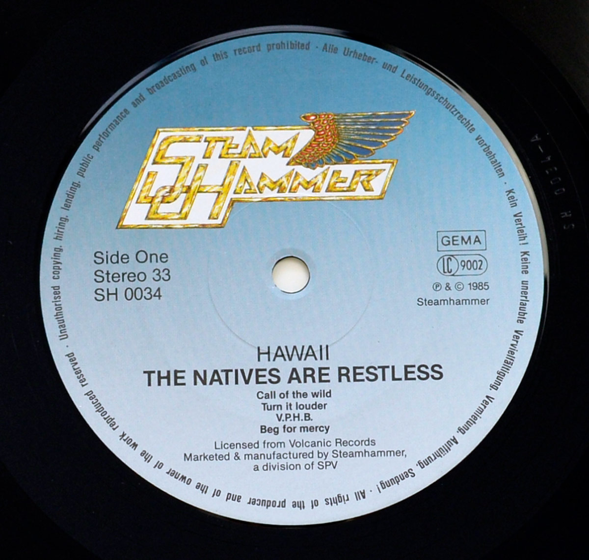 Close-up of Side One record label for the 1985 album The Natives Are Restless by Hawaii on Steamhammer. Silver-blue gradient label with the gold Steamhammer winged logo at top; text includes Side One, Stereo 33, SH 0034, GEMA, LC 9002, © 1985 Steamhammer. Center spindle hole and concentric pressing rings are visible. Track list: Call of the wild; Turn it louder; V.P.H.B.; Beg for mercy. Footer: Licensed from Volcanic Records; marketed & manufactured by Steamhammer, a division of SPV; legal rim text around the edge.