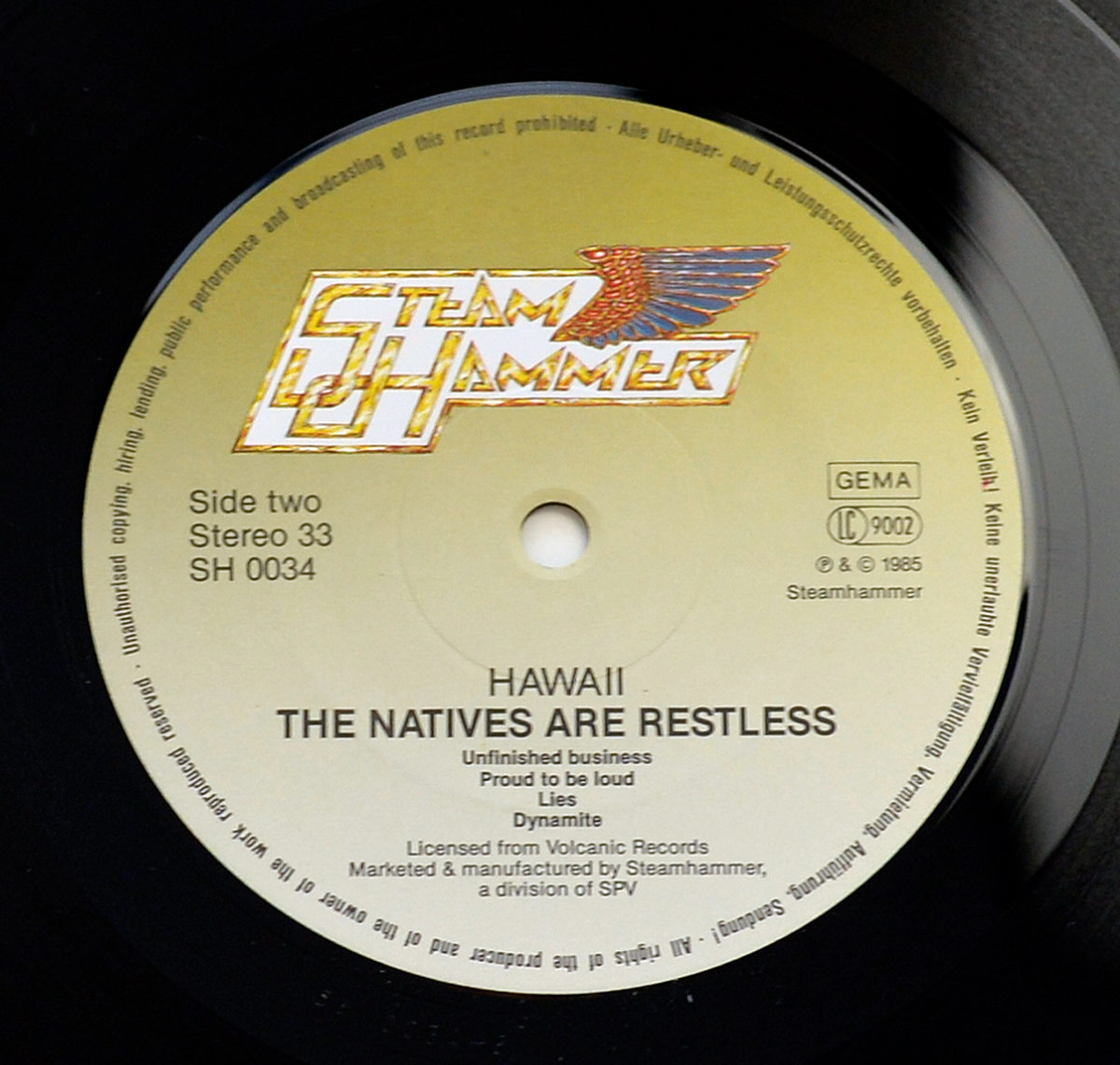 Close-up of Side Two record label for Hawaii’s 1985 album The Natives Are Restless on Steamhammer. Warm sand-to-gold gradient label with the winged Steamhammer logo at the top. Left block reads Side two, Stereo 33, SH 0034. Right block shows GEMA, LC 9002, and © 1985 Steamhammer. Center spindle hole and pressing rings are visible. Track list printed below the title: Unfinished business; Proud to be loud; Lies; Dynamite. Legal rim text runs around the perimeter.