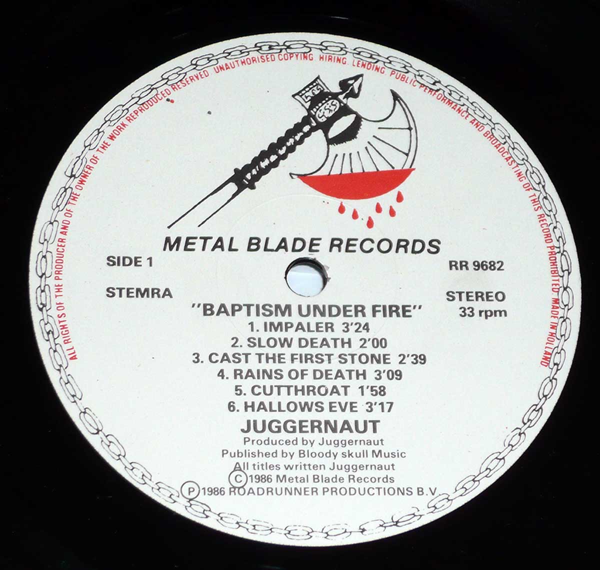 A close-up of the Side One label from Juggernaut's 'Baptism Under Fire' vinyl record, released by Metal Blade Records. The label features the iconic Metal Blade logo—an ornate battle axe dripping blood—printed in black and red. Around the edges, a chain-like border encircles the design. The tracklist is printed in bold black text, listing six songs, including 'Impaler' and 'Slow Death.' Additional details such as the catalog number RR 9682, 'STEREO' format, 33 rpm speed, and production credits are also present.