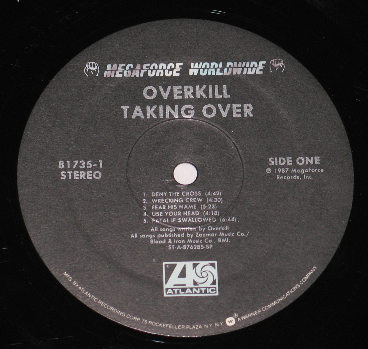 Close-up of the Side One record label of OverKill's 'Taking Over' vinyl LP, featuring a black and silver design with the Megaforce Worldwide and Atlantic Records logos at the top. The album title, band name, and tracklist for Side One are printed in bold, white capital letters. The catalog number '81735-1' and the word 'STEREO' are displayed on the left side, while the copyright information for 1987 Megaforce Records is positioned on the right.