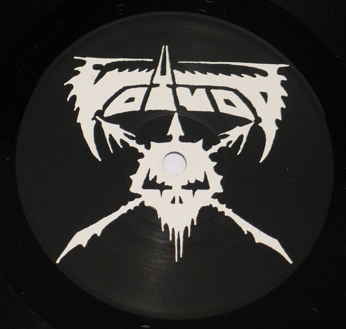 Voivod Rrröööaaarrr record label: A close-up of the record label, featuring a black background with a white, stylized Voivod logo in the center. The logo is a jagged, abstract design with the band's name, 'VOIVOD', incorporated into it. The design appears to be hand-drawn, adding to the raw and edgy feel. The center hole of the record is visible, and the grooves of the vinyl are slightly visible under the black background.
