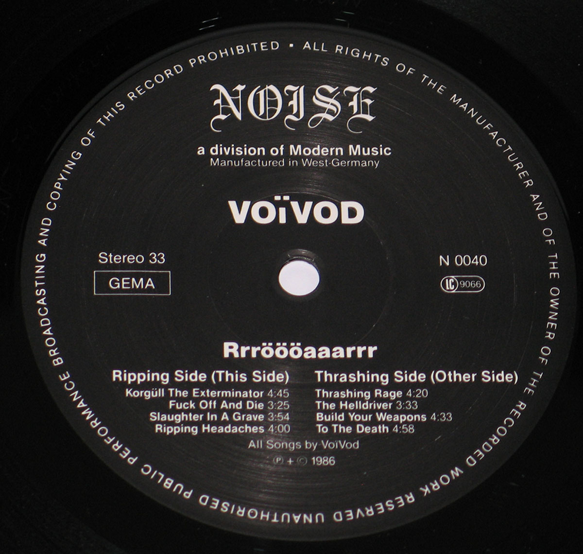 Voivod Rrröööaaarrr record label: A close-up of the record label, featuring a black background with white text. The band's name, 'VOIVOD', is prominently displayed in a stylized, bold font. The album title, 'Rrröööaaarrr', is written below in a similar font. The label also lists the song titles for both sides of the record, 'Ripping Side (This Side)' and 'Thrashing Side (Other Side)'. The 'NOISE' logo is visible at the top, indicating the record label. The label also includes details like 'Stereo 33', 'GEMA', and the catalog number 'N 0040'. The text is arranged in a circular pattern around the center hole of the record.