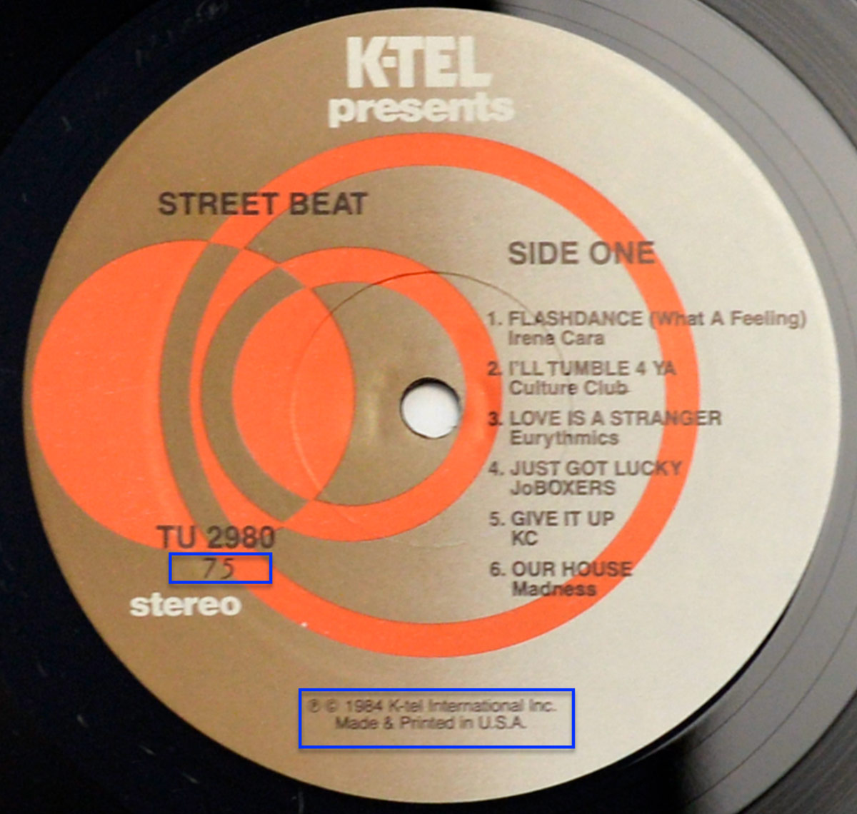 Close-up of the Side One record label of the 1984 K-Tel compilation Street Beat: New Music Hot Sounds. The label has a metallic beige background with overlapping bold orange concentric circle graphics radiating outward, giving a retro-futuristic feel. The text at the top reads K-Tel presents in white. On the left is the album title Street Beat in black, while the right side lists SIDE ONE followed by the six tracks: 1. Flashdance (What a Feeling) – Irene Cara, 2. I'll Tumble 4 Ya – Culture Club, 3. Love Is a Stranger – Eurythmics, 4. Just Got Lucky – JoBoxers, 5. Give It Up – KC, and 6. Our House – Madness. The catalog number TU 2980 is printed in bold at the bottom left above the word stereo. At the bottom edge, small text notes © 1984 K-Tel International Inc. Made & Printed in U.S.A.