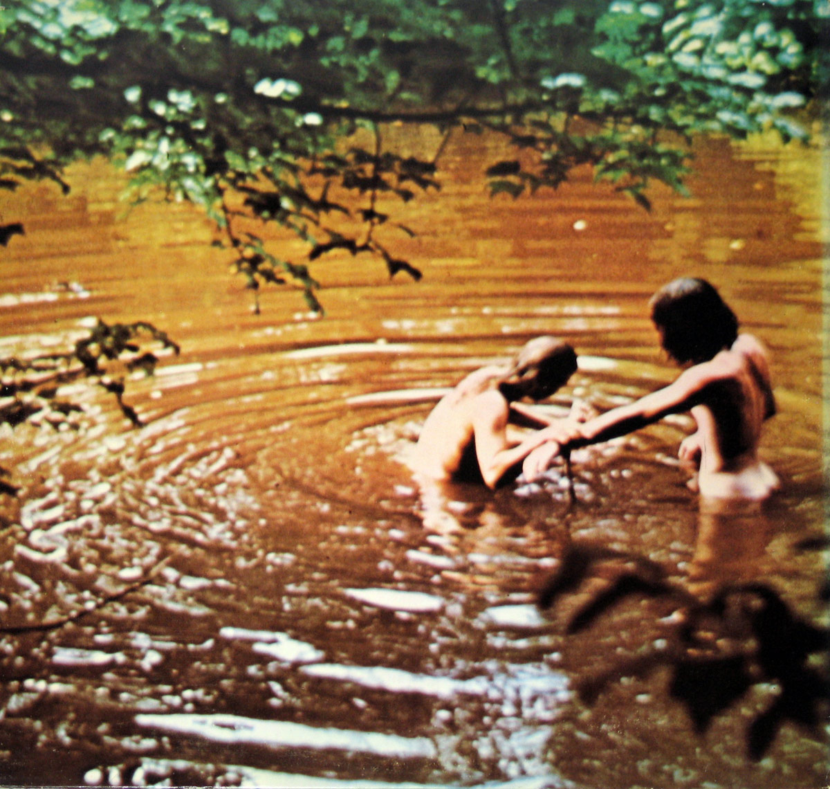 Inside sleeve photo of Woodstock Music from the Original Soundtrack USA, showing two nude individuals bathing in a pond surrounded by lush green foliage, with ripples forming on the water’s surface.